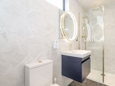 A bathroom with a wall-mounted sink and illuminated round mirror next to a toilet and a glass shower enclosure at Bluebell Cabin in Ullswater