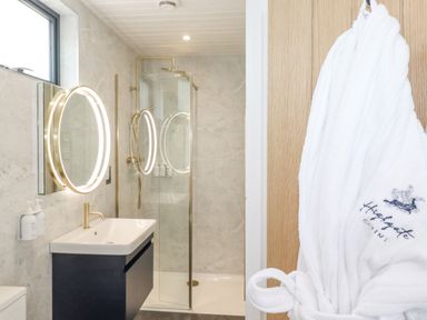 A bathroom with a sink vanity a round illuminated mirror a shower with glass door and a white bathrobe hanging on the door at Bluebell Cabin in Ullswater