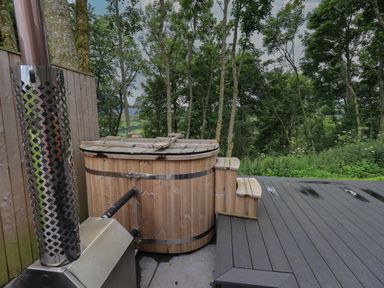 A wooden hot tub with a cover and metal stove on a deck overlooking trees and greenery at Bluebell Cabin in Ullswater
