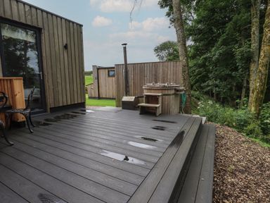 An outdoor wooden deck with a hot tub and black metal chairs surrounded by trees at Bluebell Cabin in Ullswater