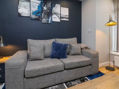 A living room with a gray sofa with cushions a side table with a table lamp a floor lamp and abstract paintings on a dark wall at The Dollhouse in Cayton Bay