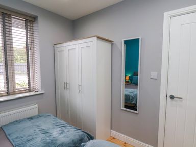 A bedroom corner with a bed covered with a blue blanket a white wardrobe a wall mirror and a window with blinds at The Dollhouse in Cayton Bay