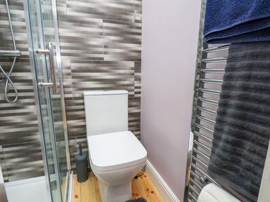 A bathroom with a toilet, glass shower enclosure, striped tiled wall, towel radiator with towels, and a wooden floor at The Dollhouse in Cayton Bay