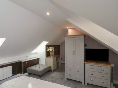 An attic bedroom with slanted ceilings featuring a wardrobe, dresser with a television, a desk with flowers, a cushioned bench, and a bed at The Dollhouse in Cayton Bay