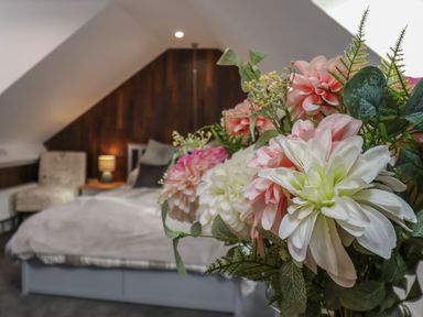 A bedroom with a bed bedside table and a chair with flowers in the foreground at The Dollhouse in Cayton Bay