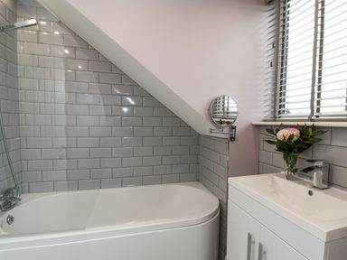 A bathroom with a bathtub shower, tiled walls, a sink cabinet, a round wall mirror, and flowers on the sink at The Dollhouse in Cayton Bay