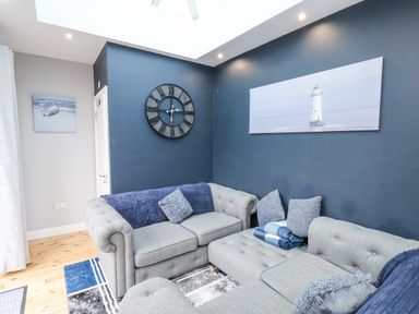 A sitting room with a grey sectional sofa a large wall clock and paintings on blue and grey walls at The Dollhouse in Cayton Bay