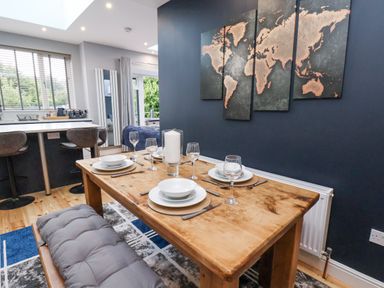 A dining area with a wooden table set with plates and glasses a bench with cushion a world map wall art and a kitchen counter with stools at The Dollhouse in Cayton Bay