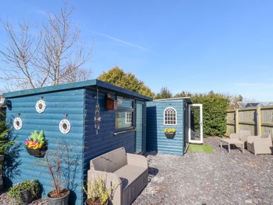An outdoor area with blue wooden sheds beige seating and potted plants at The Dollhouse in Cayton Bay