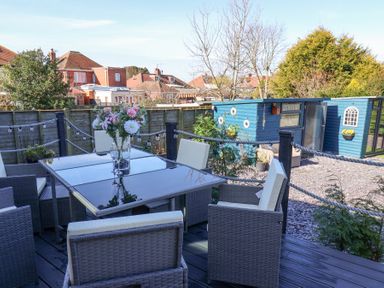 An outdoor dining area with a glass table and cushioned chairs next to a blue garden shed at The Dollhouse in Cayton Bay