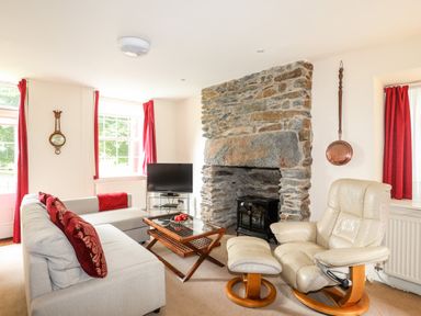 A living room with a stone fireplace a beige armchair a light grey sofa and red curtains at Ty Nain in Beddgelert