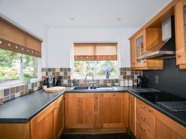 A kitchen with wooden cabinets a black countertop a stainless steel sink windows with brown blinds and various kitchen items at Ty Nain in Beddgelert