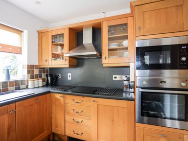 A kitchen with wooden cabinets a black countertop a built in oven and microwave a stainless steel extractor hood and a window with a brown roller blind at Ty Nain in Beddgelert