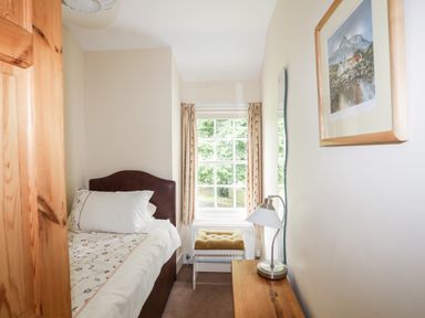A small bedroom with a single bed wooden bedside table with lamp window with beige curtains and a cushioned stool at Ty Nain in Beddgelert