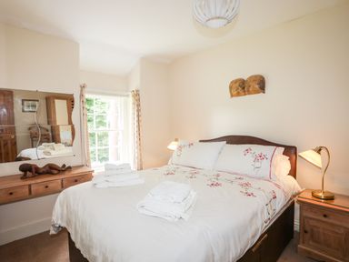 A bedroom with a double bed with floral bedding towels on the bed a wooden bedside table with a lamp a wooden dresser with a mirror and a window with curtains at Ty Nain in Beddgelert