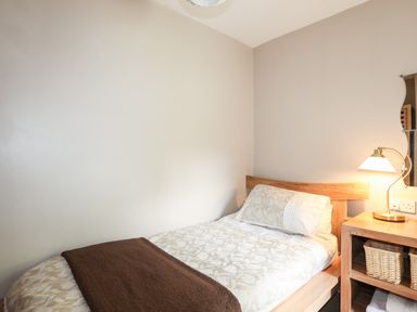 A single bed with patterned bedding and a brown throw next to a wooden bedside table with a lamp and baskets at Ty Nain in Beddgelert