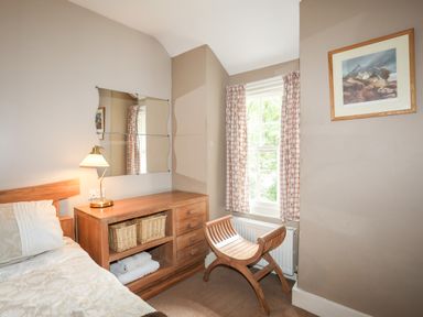A bedroom corner with a bed, wooden dresser with baskets, a lamp, a window with curtains, a wooden chair, and a wall painting at Ty Nain in Beddgelert