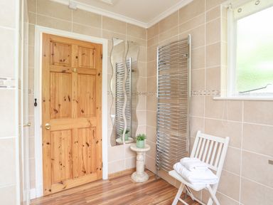 A tiled bathroom with a wooden door a metal towel radiator a small table with a plant a white chair with folded towels and a window at Ty Nain in Beddgelert