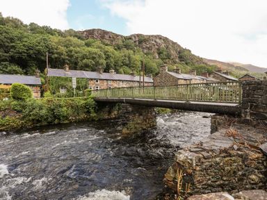 A stone bridge over a river with stone houses and a hillside in the background at Ty Nain in Beddgelert