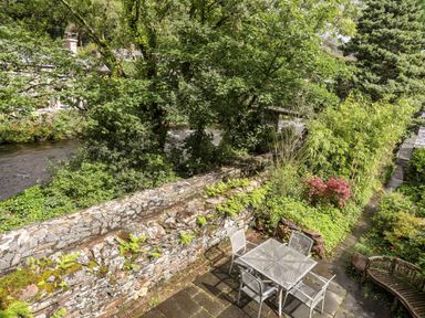 A garden patio with a metal table and four chairs surrounded by greenery and a stone wall at Ty Nain in Beddgelert