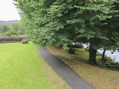 A paved path curving beside a tree and a river with a green lawn and a flower planter at Ty Nain in Beddgelert