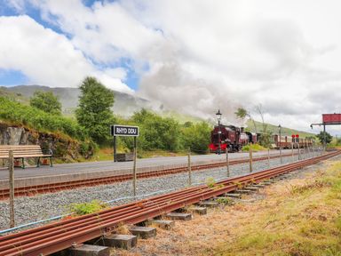 A steam train on tracks near a platform with a sign that reads Rhyd Ddu in a rural area