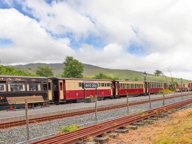 A train with red and cream carriages at Rhyd Ddu station with tracks and mountains in the background at Ty Nain in Beddgelert
