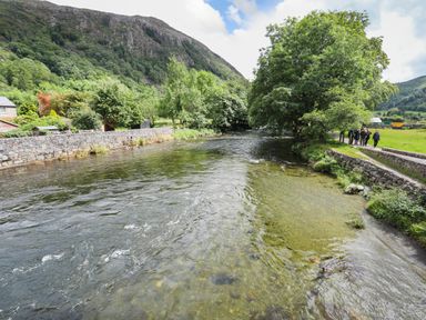 A river with stone walls on both sides and trees along the banks with people walking on a path at Ty Nain in Beddgelert