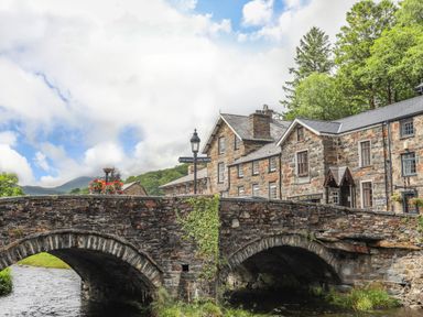 A stone bridge over a river with stone buildings and trees in the background at Ty Nain Beddgelert