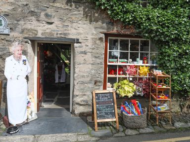 An exterior view of a stone building with an open doorway and a window displaying colorful items a cutout figure of a woman with a dog near the entrance and a chalkboard sign advertising fudge at Ty Nain in Beddgelert