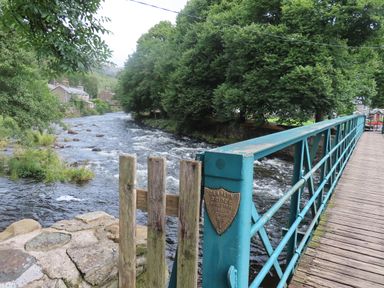 A turquoise metal pedestrian bridge over a flowing river with trees and houses in the background at Ty Nain in Beddgelert