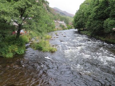 A river flowing through a village with houses trees and hills in the background at Ty Nain in Beddgelert