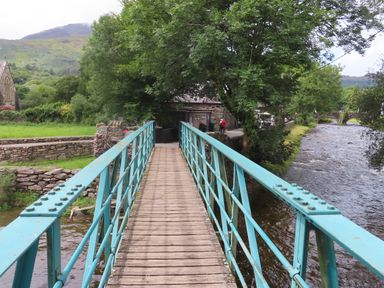 A wooden footbridge with blue railings over a river beside a stone wall and trees at Ty Nain in Beddgelert