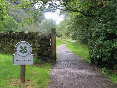 A gravel path next to a mossy stone wall and a National Trust sign for Aberglaslyn surrounded by trees and greenery