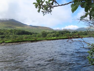 A river flowing with grassy banks and trees under a partly cloudy sky with hills in the background at Ty Nain in Beddgelert