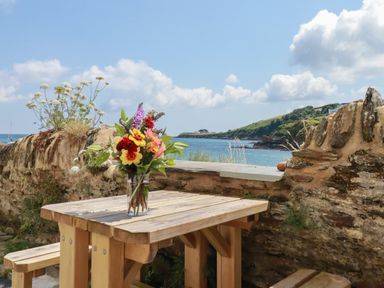 A wooden picnic table with a vase of flowers near a stone wall overlooking a coastal view at Chy Rani in Portmellon near Mevagissey