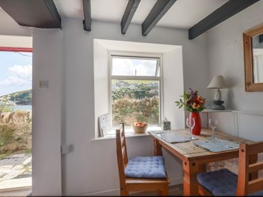 A dining area with a wooden table and chairs with blue cushions a flower vase and window showing outdoor view at Chy Rani in Portmellon near Mevagissey