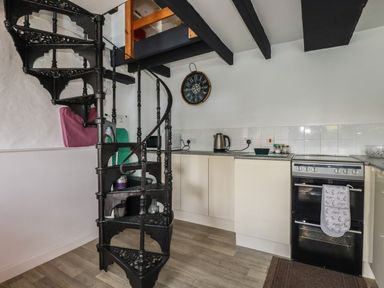 A kitchen with a black spiral staircase a black oven cream cabinets a kettle on the countertop and a wall clock at Chy Rani in Portmellon near Mevagissey