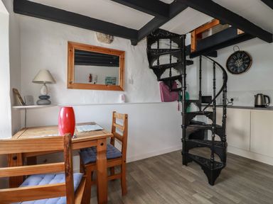 A dining area with a wooden table and chairs a red vase a table lamp a mirror on the wall a black spiral staircase and a kitchen counter with a kettle at Chy Rani in Portmellon near Mevagissey