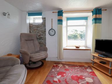 A sitting room with two armchairs a wall clock a window with curtains a television on a stand and a patterned rug at Chy Rani in Portmellon near Mevagissey