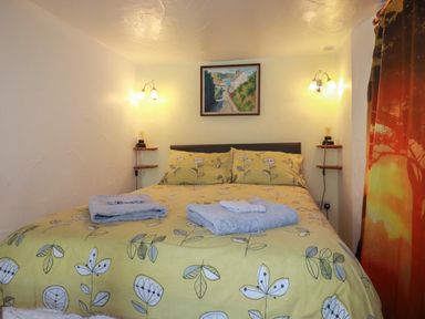 A bedroom with a bed with yellow floral bedding two folded towels wall lights a painting and a curtain with a sunset scene at Chy Rani in Portmellon near Mevagissey