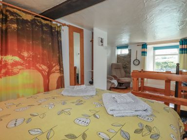A bedroom with a bed covered with yellow leaf patterned bedding and folded towels on top a curtain with a tree design a chair a wall clock and a window at Chy Rani Portmellon near Mevagissey