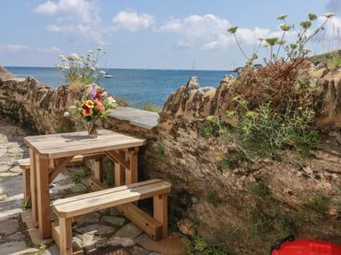 A wooden picnic table with a flower vase on a stone patio next to a stone wall overlooking the sea at Chy Rani in Portmellon near Mevagissey