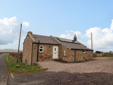 A stone cottage with solar panels on the roof near a country road at Henhill Lodge in Chathill near Seahouses