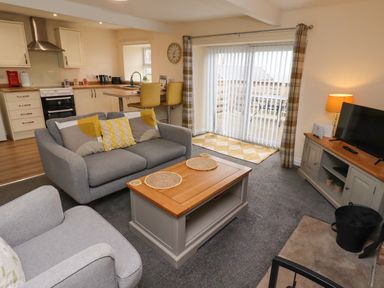 An open plan living room and kitchen with grey sofas a wooden coffee table a TV on a cabinet and a kitchen area with stools and cabinets at Henhill Lodge in Chathill near Seahouses
