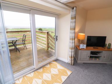 A room with sliding glass door leading to a wooden deck with table and chair and a cabinet with TV and lamp at Henhill Lodge in Chathill near Seahouses