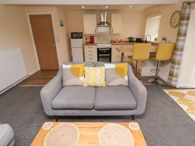 A living area with a gray sofa with yellow and gray cushions a wooden coffee table with circular woven mats a small kitchen with white cabinets a stove a refrigerator a microwave and two yellow bar stools at Henhill Lodge in Chathill near Seahouses