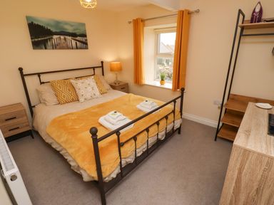 A bedroom with a metal bed frame and yellow bedding two bedside tables a window with yellow curtains and a wooden shelving unit at Henhill Lodge in Chathill near Seahouses