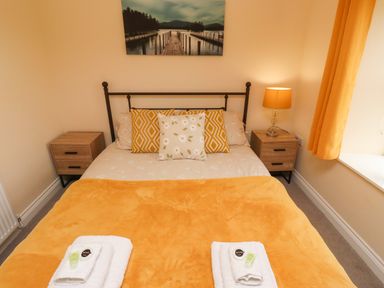 A bedroom with a double bed with yellow and floral pillows two bedside tables with a lamp and a window with yellow curtains at Henhill Lodge in Chathill near Seahouses