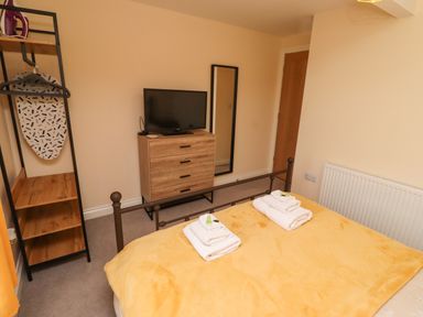 A bedroom with a bed covered by a yellow blanket with folded towels on top a wooden dresser with a television a standing mirror and an open wardrobe with an ironing board at Henhill Lodge in Chathill near Seahouses